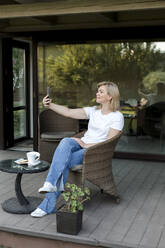 Lächelnde Frau, die ein Selfie mit ihrem Smartphone macht und auf einem Stuhl auf der Veranda sitzt - LLUF00763