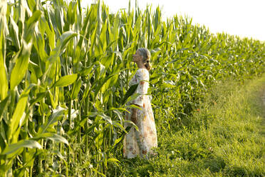 Senior woman wearing summer dress standing in front of corn field - FLLF00657