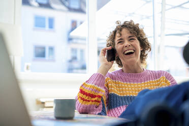 Cheerful woman talking on smart phone sitting at home - JOSEF11642