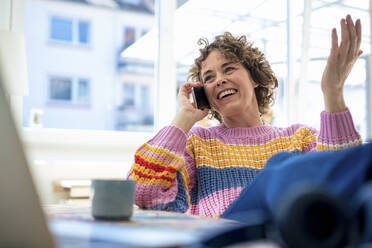 Smiling woman talking on mobile phone sitting at home - JOSEF11643