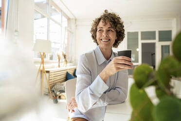 Happy businesswoman with disposable coffee cup sitting at desk in office - JOSEF11649