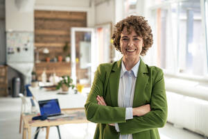 Happy mature businesswoman wearing green blazer standing with arms crossed in office - JOSEF11655