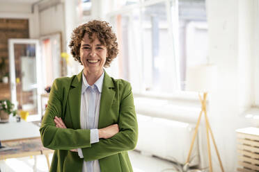 Happy businesswoman wearing green blazer standing with arms crossed in office - JOSEF11656