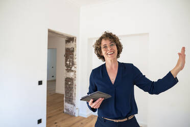 Smiling real estate agent holding tablet PC gesturing in new home - JOSEF11692