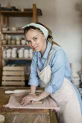 Lächelnde Kunsthandwerkerin knetet Ton auf einer Werkbank im Kunstatelier - LLUF00779