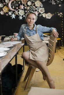 Lächelnder Kunsthandwerker auf einem Stuhl im Kunstatelier sitzend - LLUF00792