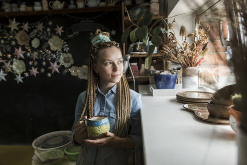 Smiling craftsperson holding ceramic mug looking through window at workshop - LLUF00800