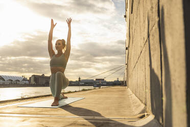 Frau mit erhobenen Armen übt Yoga auf der Promenade bei Sonnenuntergang - UUF27014