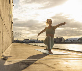 Lächelnde Frau mit ausgestreckten Armen übt Yoga auf der Promenade bei Sonnenuntergang - UUF27015