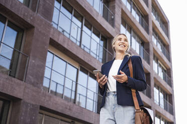 Happy woman with bag and smart phone standing in front of building - UUF27032