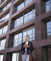 Smiling woman text messaging through mobile phone in front of building - UUF27034