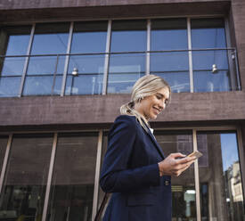 Lächelnde blonde Frau, die mit ihrem Mobiltelefon vor einem Gebäude im Internet surft - UUF27035