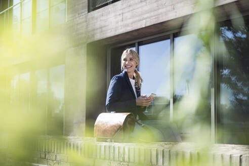 Happy young woman with smart phone sitting on wall - UUF27038