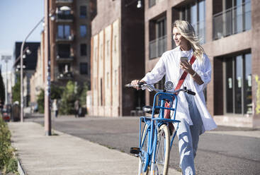 Smiling blond woman walking with bicycle on footpath - UUF27054