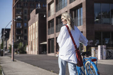 Lächelnde Frau zu Fuß mit Fahrrad vor einem Gebäude an einem sonnigen Tag - UUF27057