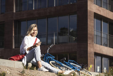 Lächelnde blonde Frau, die ein Smartphone benutzt und am Fahrrad sitzt - UUF27061