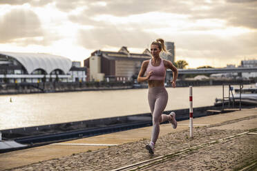 Blonde Frau joggt vor einem Fluss bei Sonnenuntergang - UUF27073