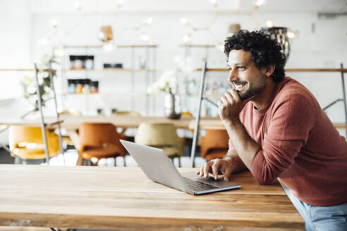 Smiling businessman with laptop on desk in office - JOSEF11724