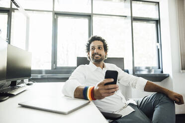 Smiling businessman with smart phone by desk at office - JOSEF11764