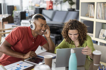 Lächelnde Geschäftsfrau, die einen Laptop mit einem Kollegen teilt, der drahtlose Kopfhörer im Büro trägt - JCICF00298