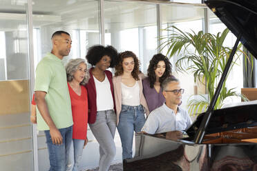 Businesswomen standing with businessman looking at colleague playing piano in office - JCICF00394