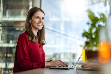 Lächelnde Geschäftsfrau mit Laptop träumt am Schreibtisch im Büro - DIGF18482