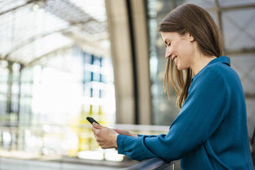 Smiling businesswoman using mobile phone in office corridor - DIGF18484