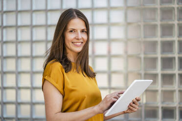 Smiling businesswoman with tablet PC by wall in office - DIGF18492