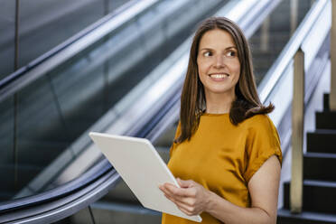 Smiling businesswoman with tablet PC by escalator - DIGF18511