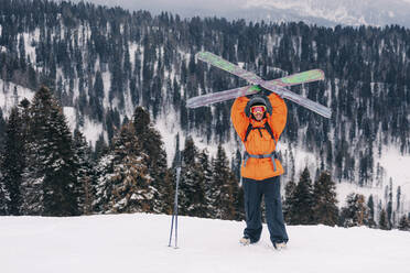 Lächelnder Skifahrer, der mit erhobenen Armen vor einem Berg steht und die Skier hält - OMIF01006