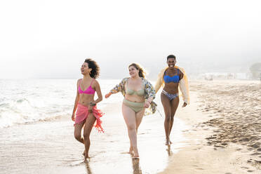 Gemischtrassige Freunde in Badekleidung, die am Strand in der Nähe des Ufers spazieren gehen - OIPF02280