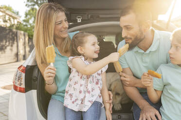 Smiling family eating ice pops sitting at back of car - JCCMF06783