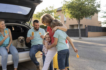 Geschwister, die ihre Großmutter umarmen, während die Eltern mit dem Hund im Kofferraum des Autos sitzen - JCCMF06793