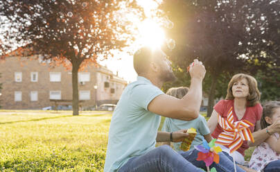 Mann bläst Seifenblasen und sitzt mit seiner Familie im Park - JCCMF06809