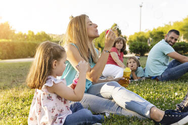 Mutter und Tochter blasen Seifenblasen und sitzen bei der Familie im Park - JCCMF06813