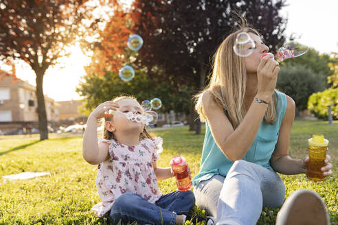 Mother and daughter blowing bubbles sitting at park - JCCMF06815