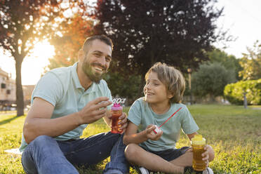 Lächelnder Vater und Sohn mit Seifenblasenstäben im Park sitzend - JCCMF06818
