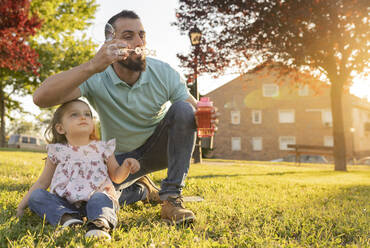 Daughter looking at father blowing bubbles at park - JCCMF06823