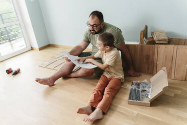 Sohn und Vater lesen Gebrauchsanweisung auf dem Boden sitzend zu Hause - OSF00534