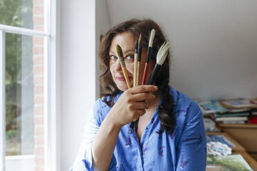 Artist holding paintbrushes by face at home - IHF01069