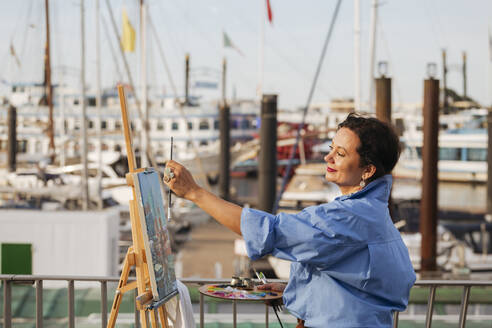 Female painter measuring by easel at harbor - IHF01076