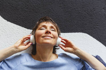 Entspannte reife Frau hört Musik an der Wand lehnend - IHF01094