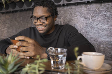 Smiling man wearing eyeglasses using smart phone in cafe - OSF00548