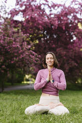 Smiling woman with hands clasped doing yoga sitting in apple blossom garden - EYAF02001