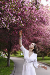 Frau mit erhobenem Arm, der nach den Blüten eines Apfelbaums im Garten greift - EYAF02012