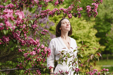 Lächelnde Frau mit geschlossenen Augen an einem Apfelblütenbaum stehend - EYAF02015