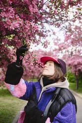 Woman wearing cap smelling apple blossoms in garden - EYAF02021
