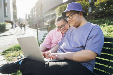 Jugendlicher benutzt Laptop bei Schwester mit Down-Syndrom auf Bank - MEUF07365
