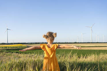 Mädchen mit ausgestreckten Armen steht in einem Feld und schaut auf Windkraftanlagen - SVKF00411
