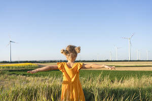 Mädchen mit ausgestreckten Armen steht in einem Feld und schaut auf Windkraftanlagen - SVKF00411
